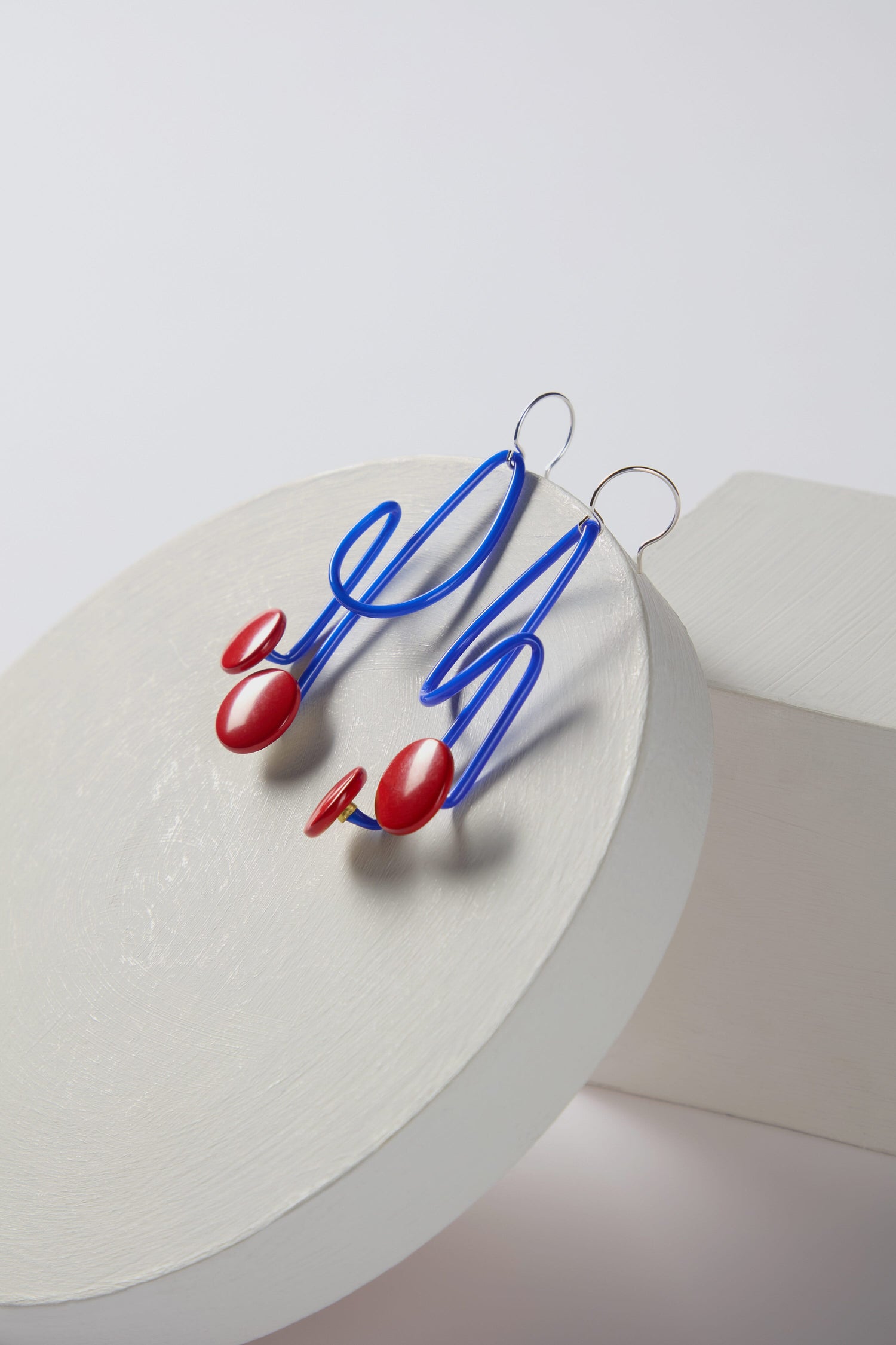 A pair of playful and bold Symphony Earrings by Samuel Coraux, featuring abstract blue wire with red circular accents, displayed on a white, round pedestal. These statement adornments are perfect for adding a pop of color to any outfit.