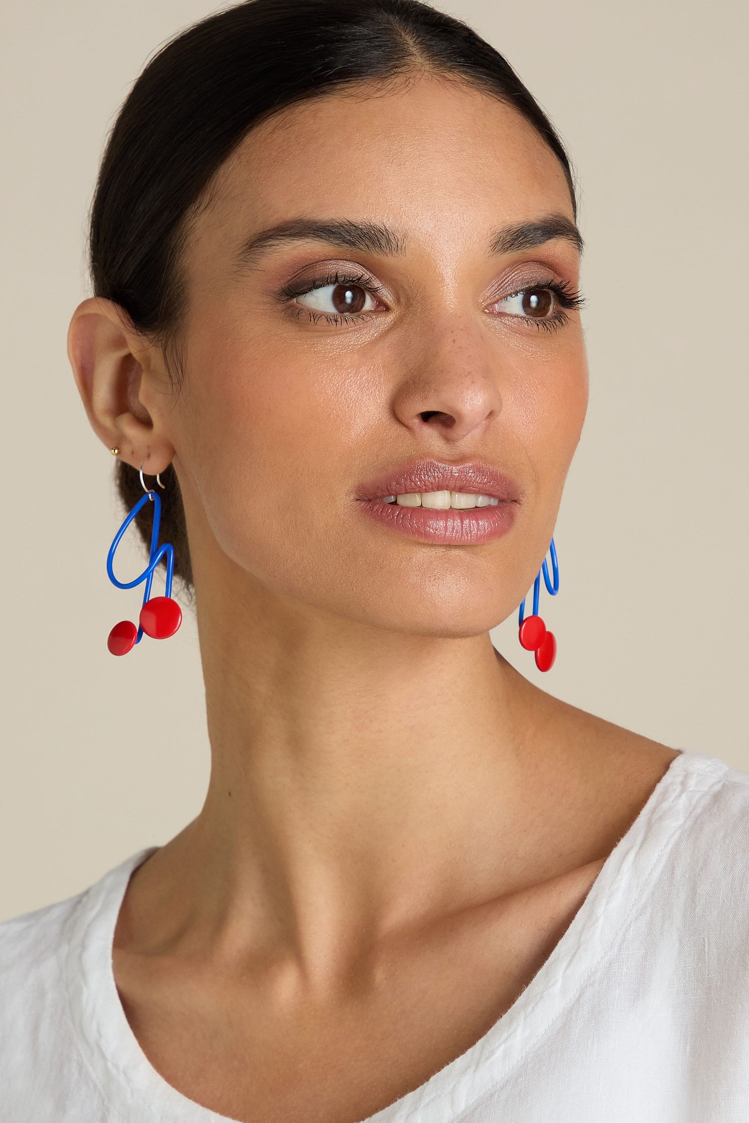 A person with dark hair is wearing a white top and playful, bold blue and red Symphony Earrings by Samuel Coraux, looking slightly to the side.