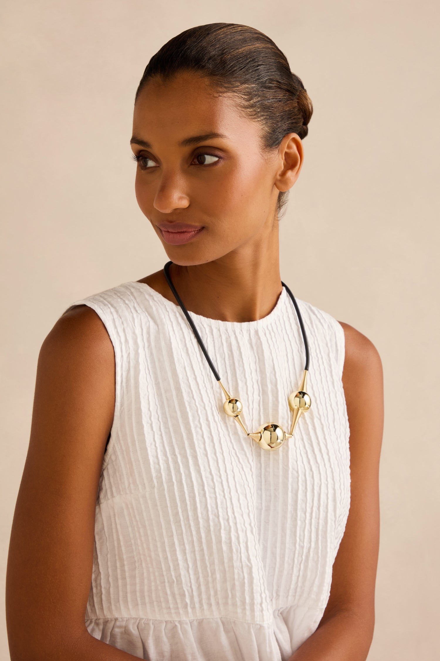 A woman looks to the side against a neutral background, wearing a sleeveless white textured dress and the Cosmos Necklace—a sculptural black silicone necklace adorned with large gold beads.