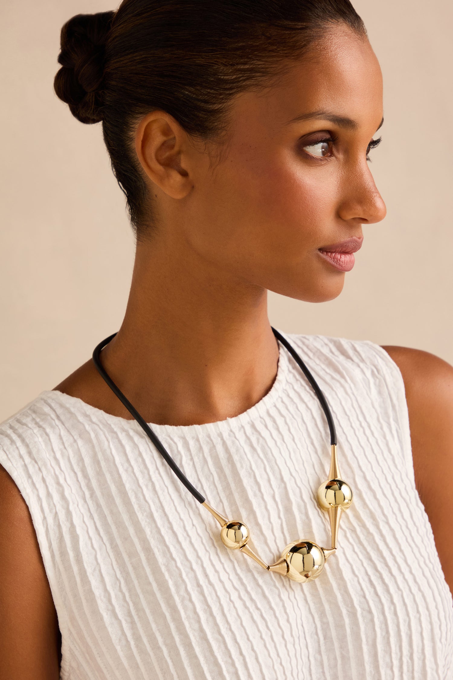 Wearing the Cosmos Necklace, a sculptural gold statement piece featuring three spherical beads and handmade in France, the woman in a white textured sleeveless top gazes to the right against a neutral background, highlighting the allure of this striking jewellery.