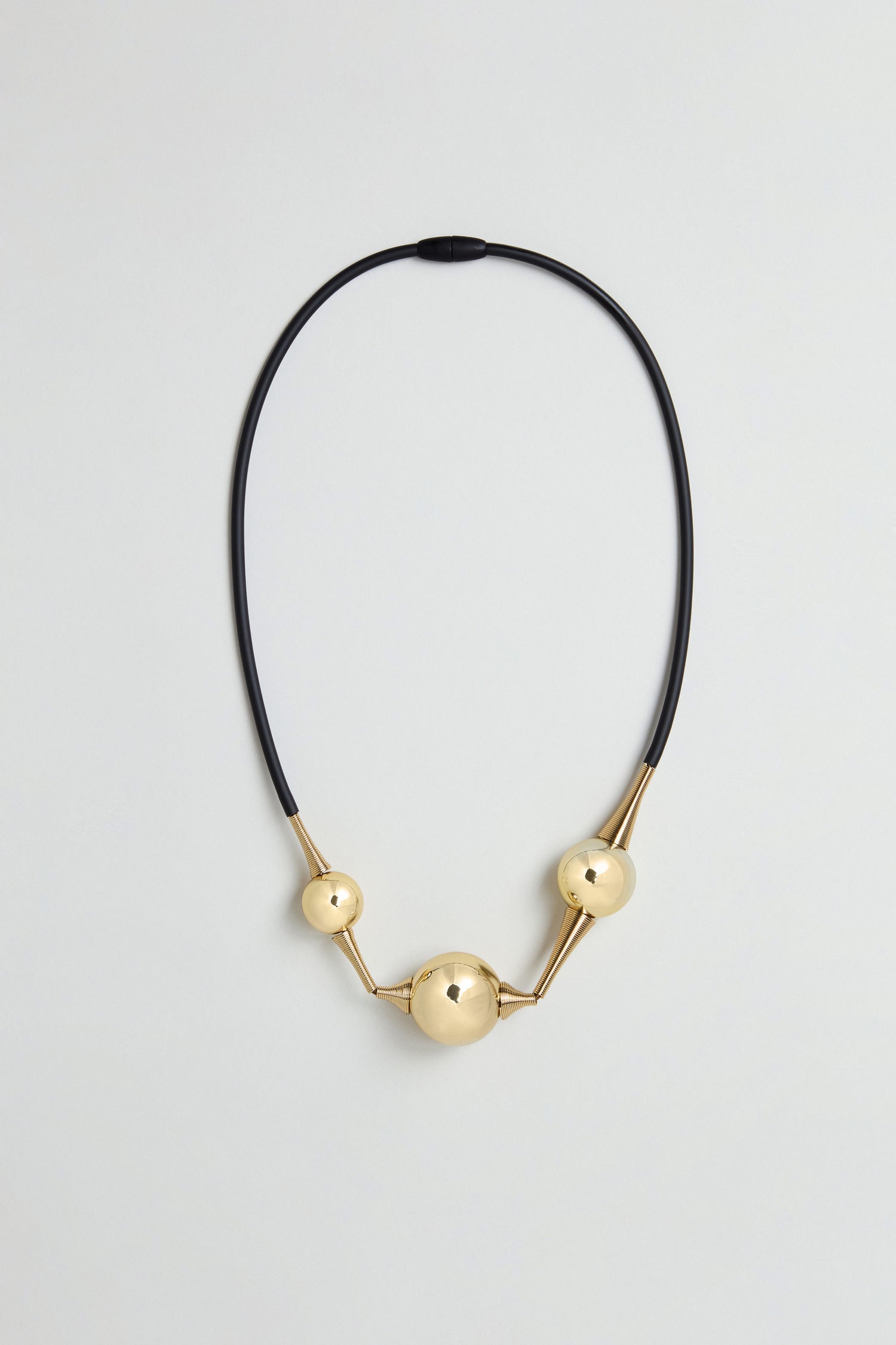 The Cosmos Necklace features three gold spherical beads of varying sizes arranged along a black silicone cord, creating a sculptural jewellery piece displayed against a plain light background.