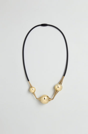 The Cosmos Necklace features three gold spherical beads of varying sizes arranged along a black silicone cord, creating a sculptural jewellery piece displayed against a plain light background.