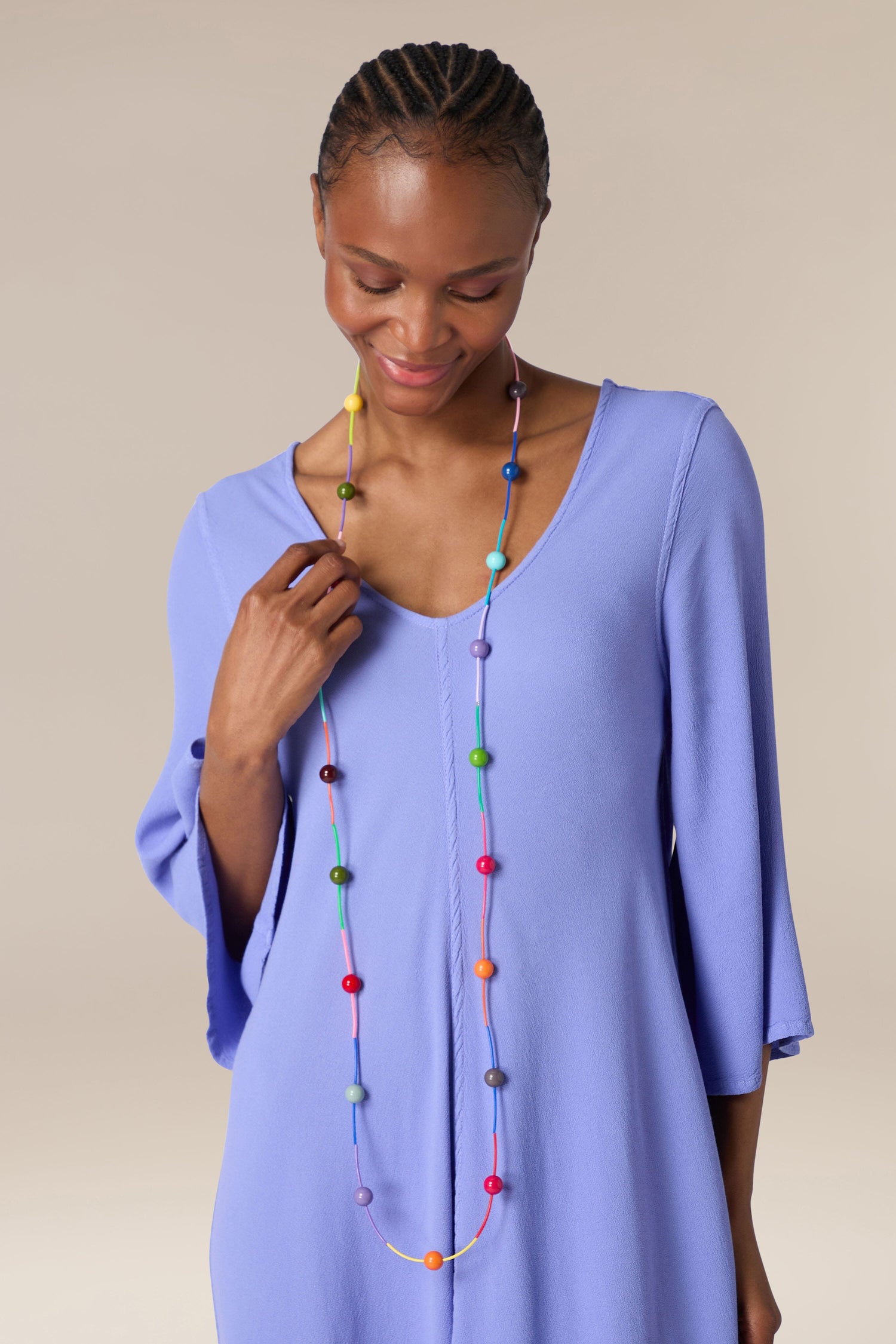 A woman in a blue top smiles gently while holding a Rainbow Sphere Colour Pop Necklace by Samuel Coraux.