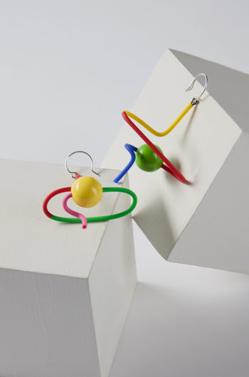 The Sketch Ball Earrings are two colorful, abstract sculpted pieces featuring twisted wire and spherical beads in yellow, green, red, and blue. Displayed on white geometric blocks, these playful accessories add a touch of drama to any outfit with their bright graphic design.