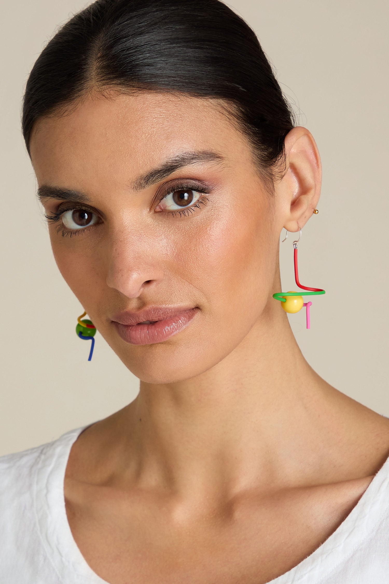 A person with long dark hair tied back, wearing Sketch Ball Earrings and a white top, looks into the camera against a neutral background.