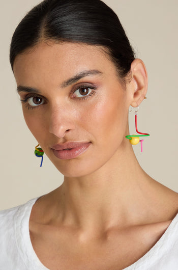 A person with long dark hair tied back, wearing Sketch Ball Earrings and a white top, looks into the camera against a neutral background.