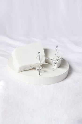 A pair of Sketch Ball Earrings by Samuel Coraux is displayed on a round white dish, with a white foam block as a backdrop, all set against a white fabric background.