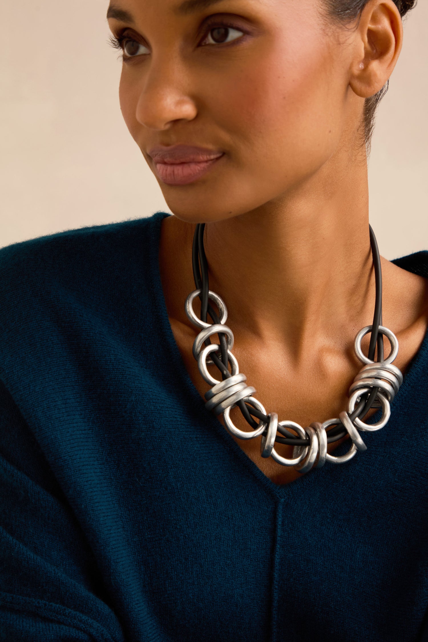 A person wears the Metal Rings Necklace—a chunky silver statement piece with a sculptural design—paired with a dark blue V-neck top, looking slightly to the side against a plain background.