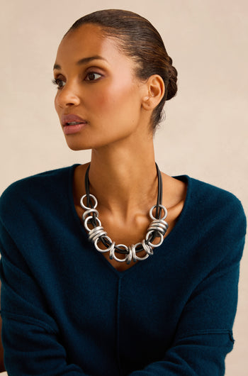 A woman with pulled-back hair wears the Metal Rings Necklace—a chunky silver piece, handmade in France—paired with a dark teal sweater, as she gazes slightly to the side against a neutral background.