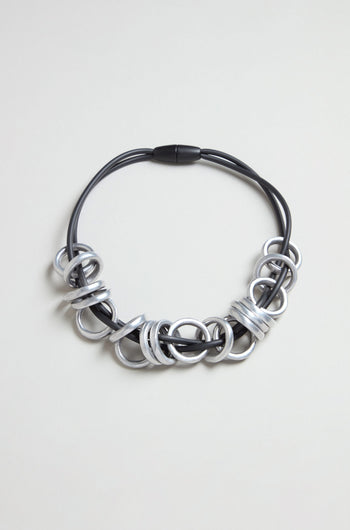 The Metal Rings Necklace features a black cord adorned with multiple large, intertwined metallic silver rings grouped at the front in a striking sculptural design, elegantly displayed against a neutral background.