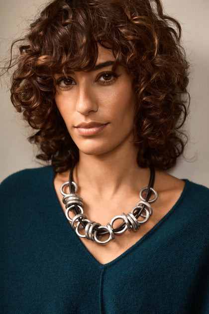 A woman with curly brown hair wears a teal V-neck sweater and the Metal Rings Necklace—a chunky, sculptural piece handmade in France—looking directly at the camera with a neutral expression.