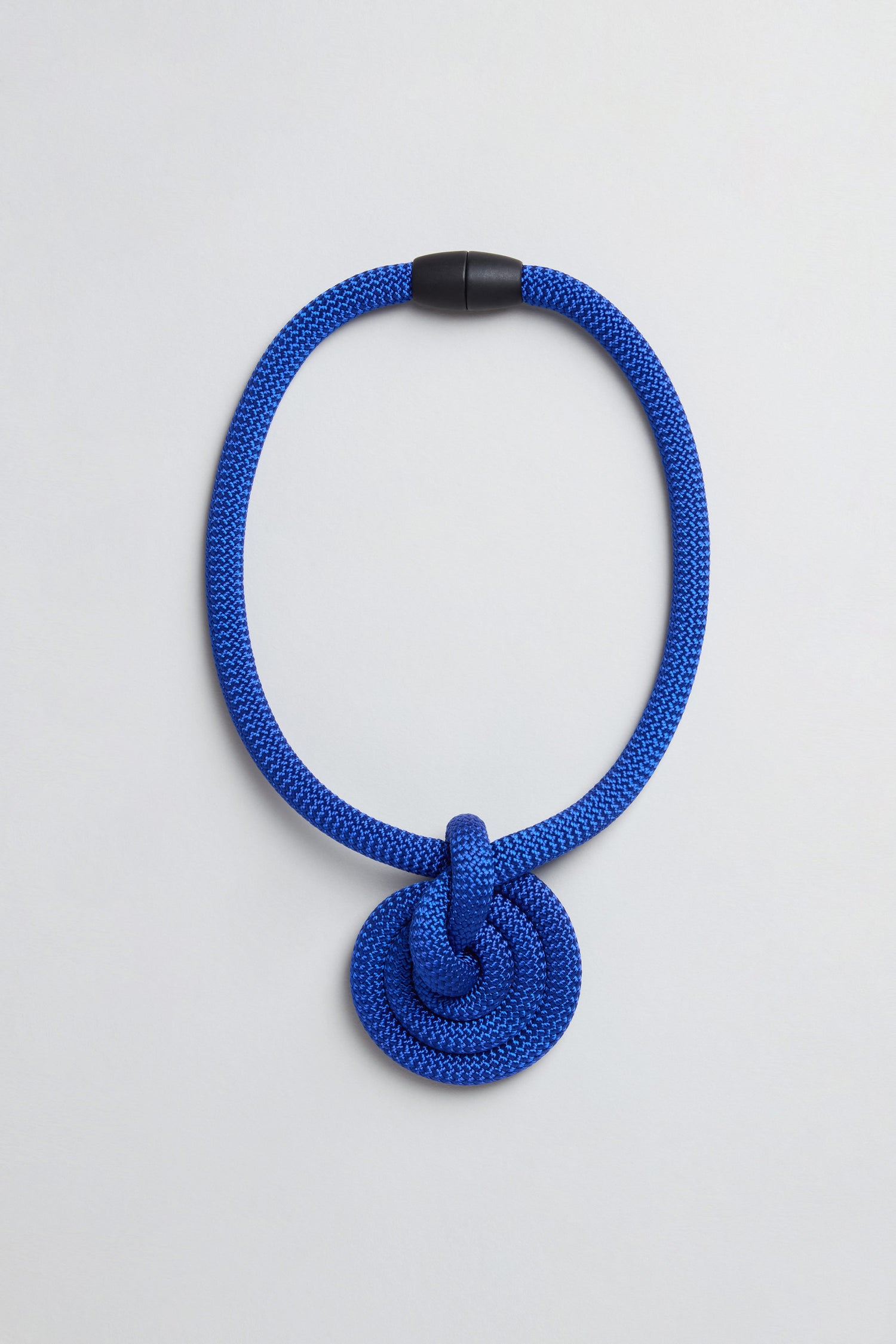 The Spiral Cord Necklace features a blue spiral cord with a large knot pendant and black clasp, handmade in Paris. Displayed on a light gray background.