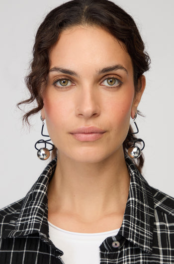 A woman with wavy brown hair, dressed in a black-and-white plaid shirt and white top, wears Graphic Squiggle Earrings and looks directly at the camera against a plain background.