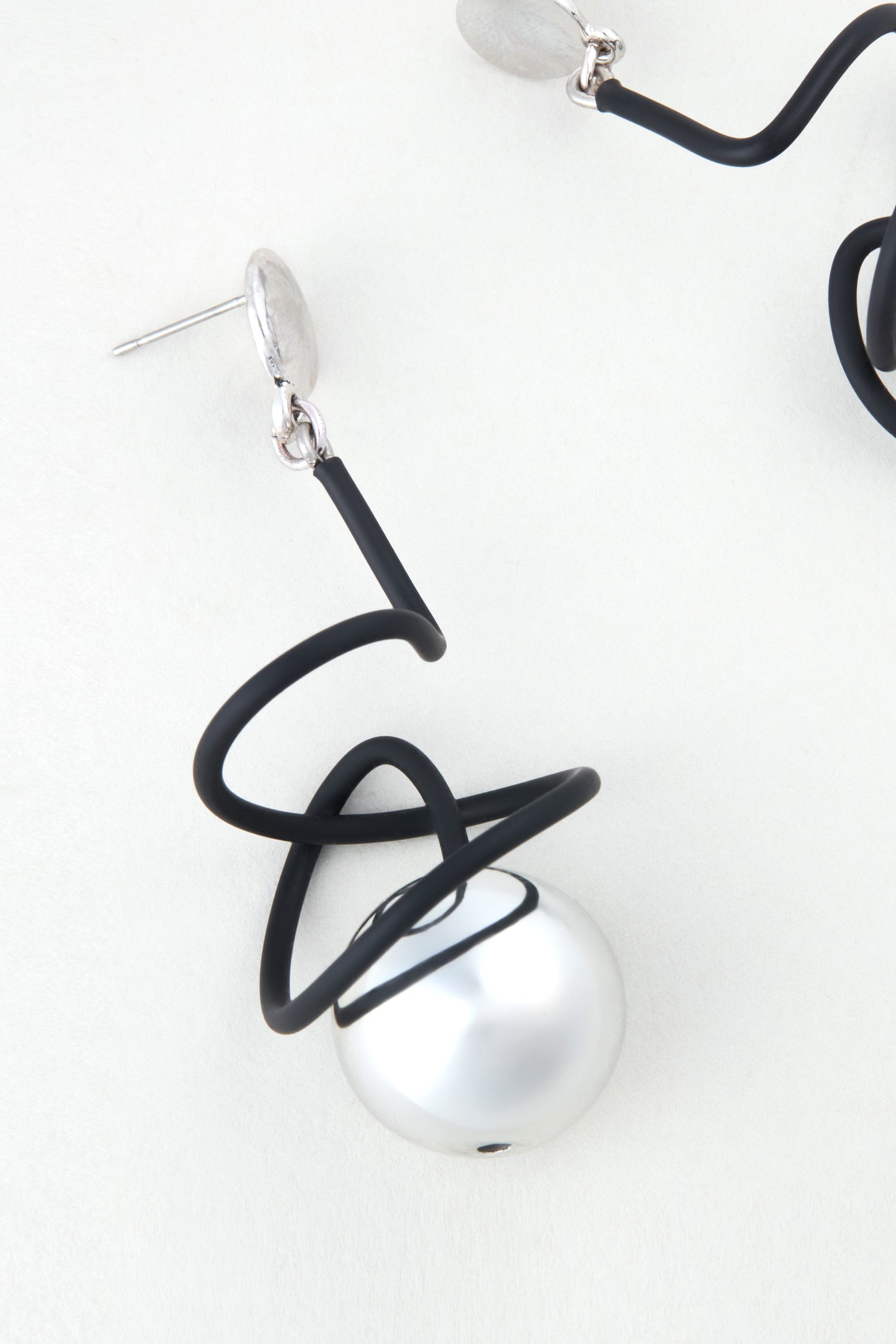 The Graphic Squiggle Earrings showcase a large round pearl wrapped in a black wire spiral, reflecting sculptural design inspired by Samuel Coraux, set against a light background.