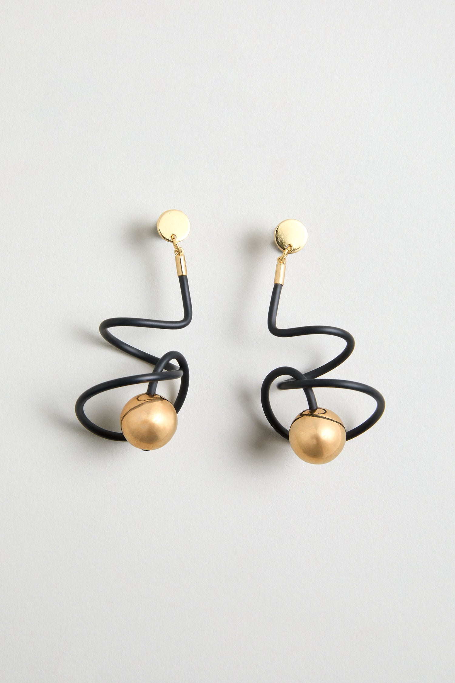 The Graphic Squiggle Earrings feature gold circular studs, black wavy wire loops, and gold sphere pendants at each end, shown on a simple light background.