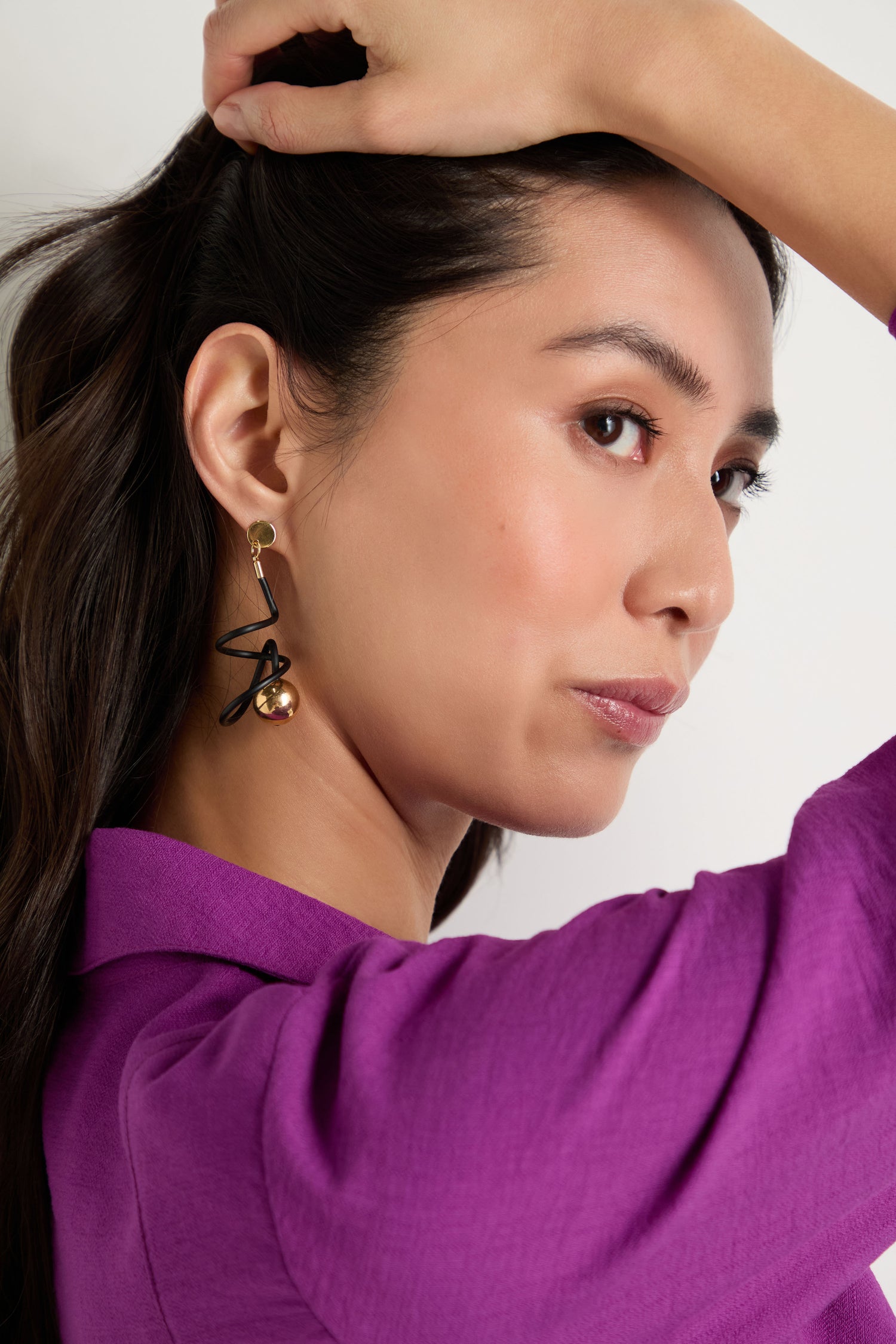 A woman in a purple top poses with one arm raised, partially in profile against a plain background, showcasing the contemporary Graphic Squiggle Earrings.
