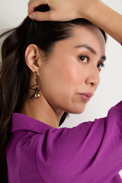 A woman in a purple top poses with one arm raised, partially in profile against a plain background, showcasing the contemporary Graphic Squiggle Earrings.
