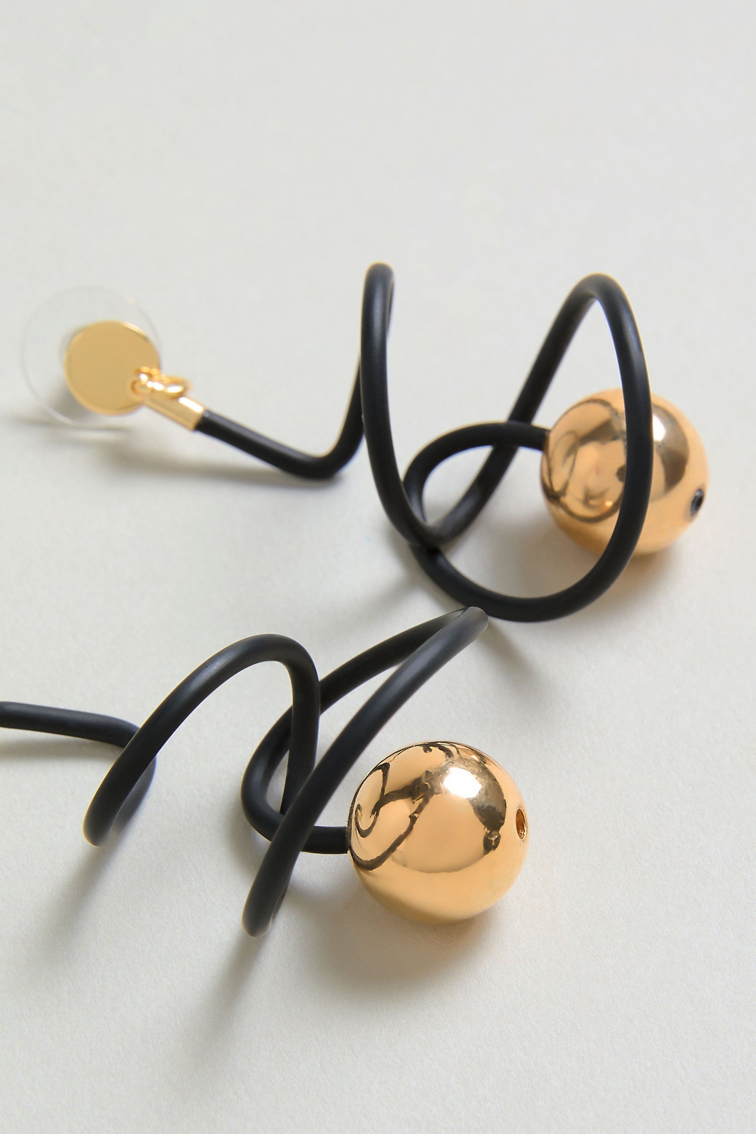 The Graphic Squiggle Earrings feature a black spiral wire with two shiny gold spheres and a clear round end, creating bold statement drop earrings shown on a light-colored surface.