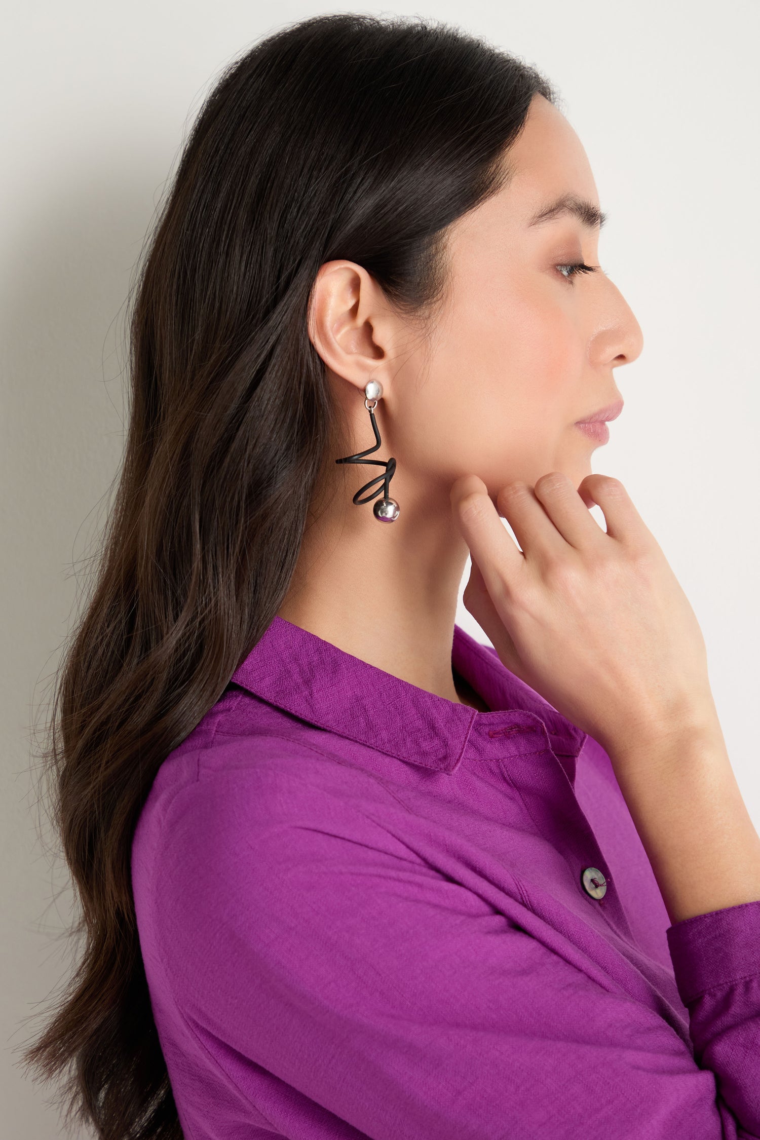 A woman in a purple shirt with long dark hair, shown in profile, thoughtfully touches her chin while wearing contemporary Graphic Squiggle Earrings featuring a bold zigzag design and pearl accents.