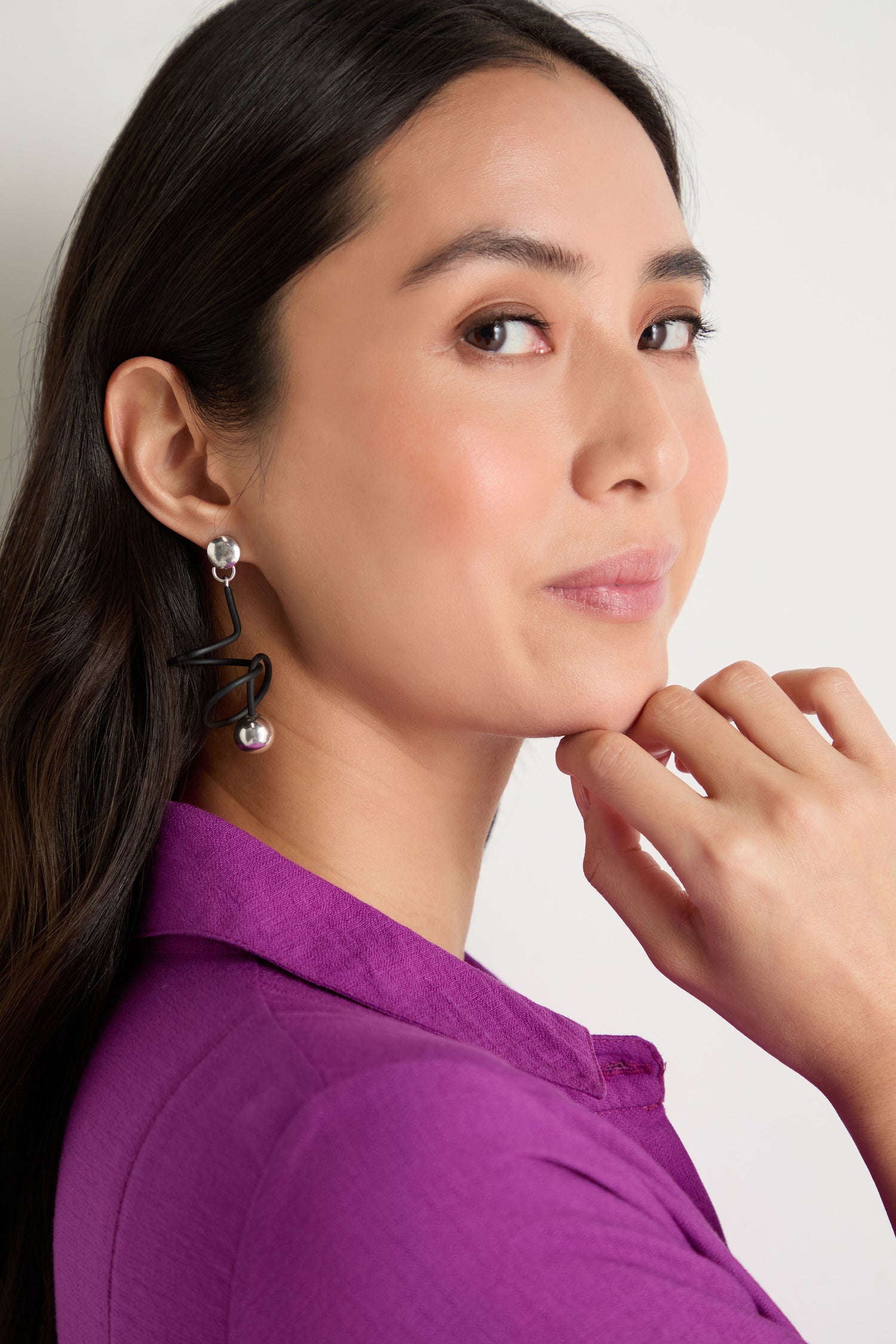 A woman with long dark hair, in a purple collared top and modern jewellery, poses with her hand near her chin to highlight her Graphic Squiggle Earrings against a plain background.