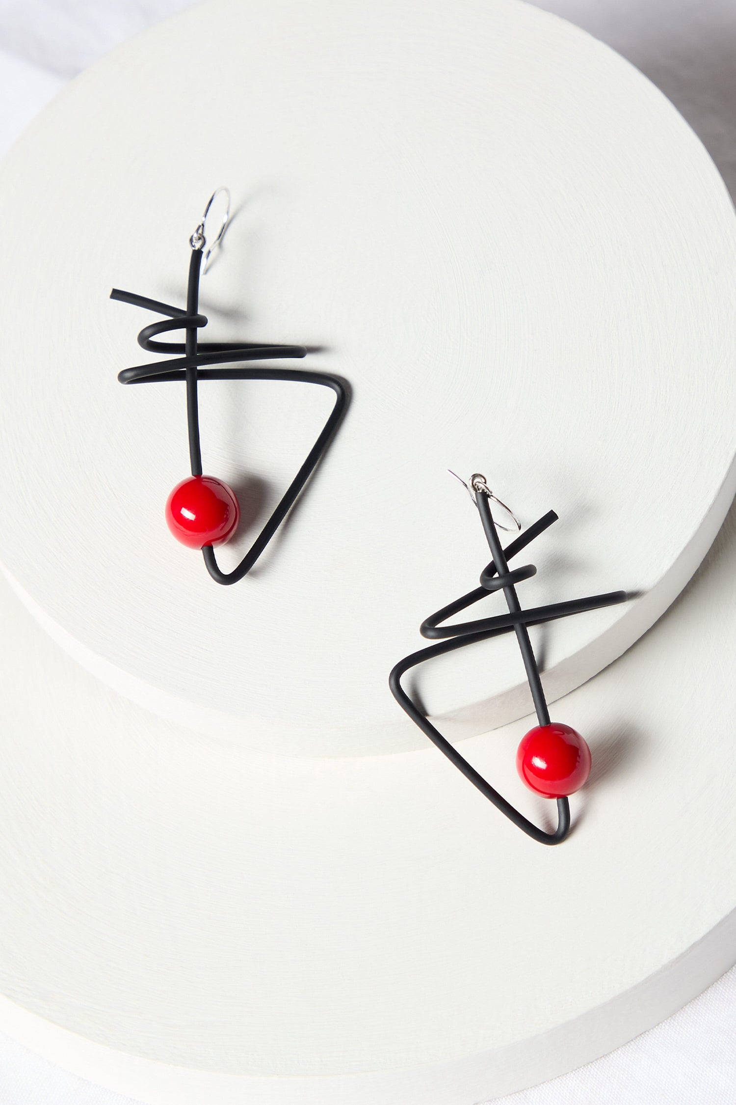 A pair of Doodle Earrings, featuring a modern abstract design with black wire and red spherical beads, crafted by renowned French jewelry maker Samuel Coraux, elegantly displayed on a white circular platform.