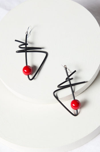 A pair of Doodle Earrings, featuring a modern abstract design with black wire and red spherical beads, crafted by renowned French jewelry maker Samuel Coraux, elegantly displayed on a white circular platform.