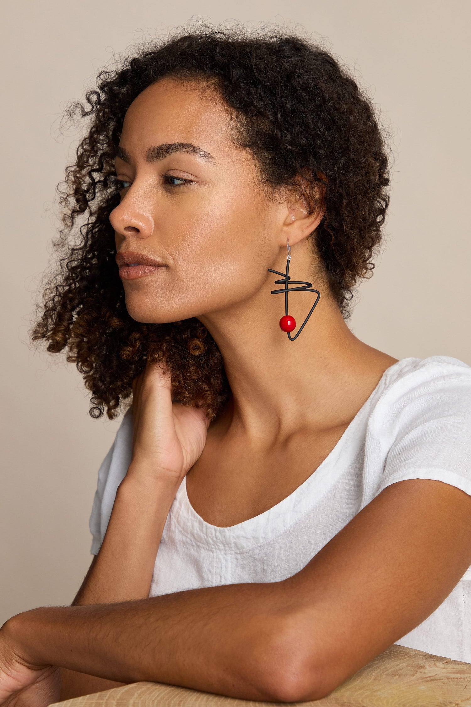 A person with curly hair wearing a white shirt and Doodle Earrings by Samuel Coraux in red and black abstract design rests their chin on their hand.