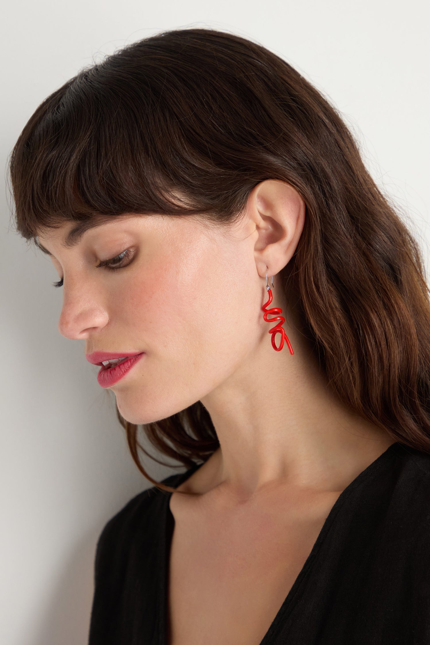 A woman with long brown hair and red lipstick wears a black top and Mini Sketch Earrings—eye-catching, red, abstract accessories—while looking down against a plain background.