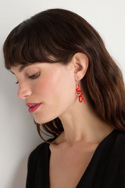 A woman with long brown hair and red lipstick wears a black top and Mini Sketch Earrings—eye-catching, red, abstract accessories—while looking down against a plain background.