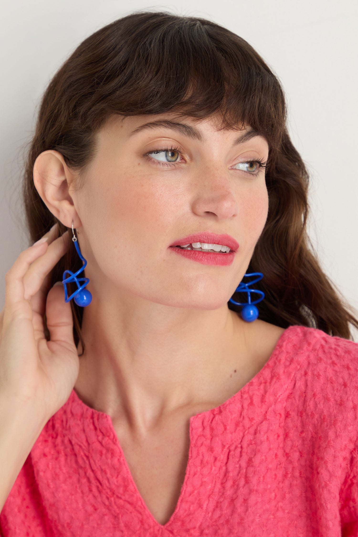 A woman in a textured pink top wears the Scribble Sphere Earrings, featuring a bold sculptural design, as she gazes sideways and touches one earring.