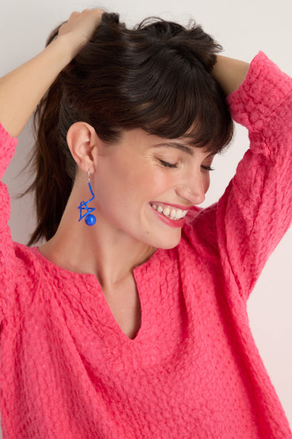 A woman smiles with eyes closed, wearing a textured bright pink top and bold Scribble Sphere Earrings—celebrated for their sculptural look and handmade in Paris—with arms raised behind her head against a plain background.