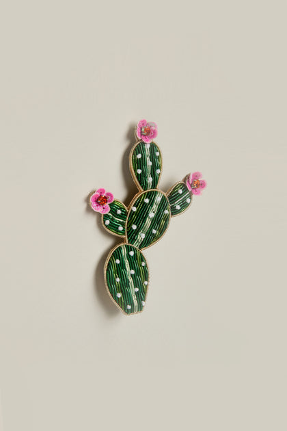 The Cactus Brooch showcases a lively burst of color with its cactus shape featuring green and white stripes, accented by three pink flowers.