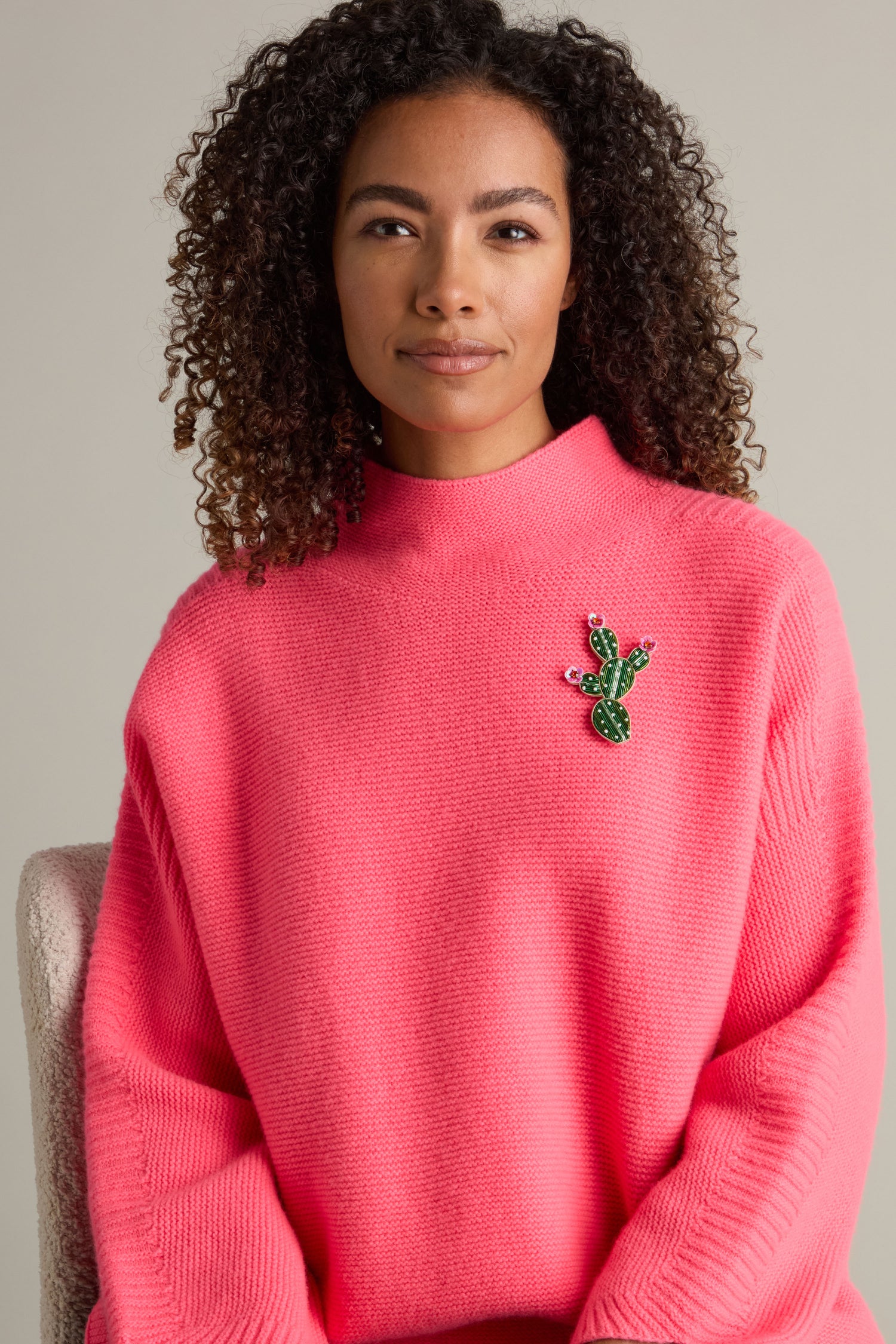 A woman with curly hair wears a bright pink sweater adorned with the vibrant "Cactus Brooch," seated against a neutral background.