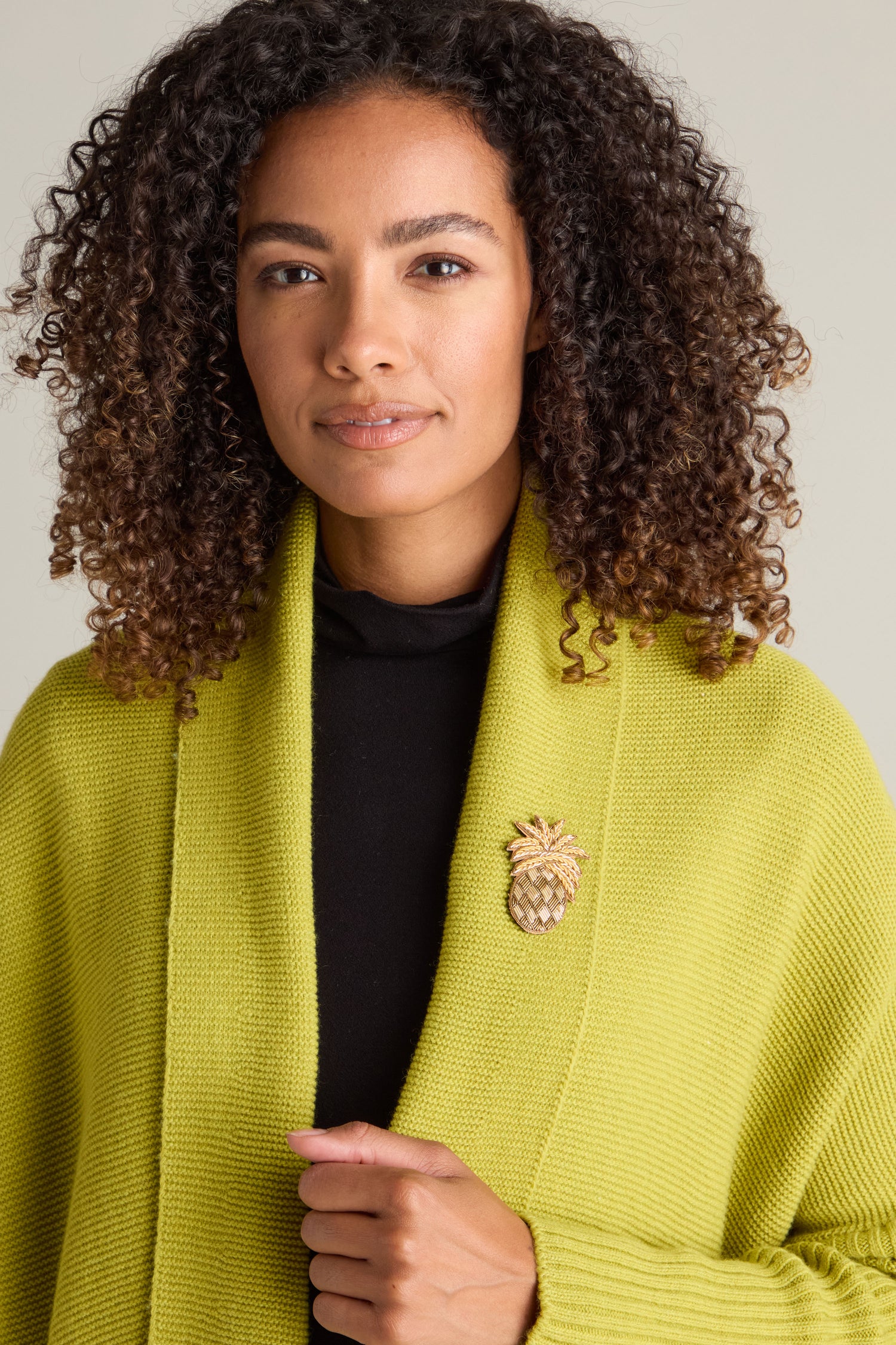 A person with curly hair wears a lime green shawl over a black shirt. The Pineapple Brooch, adorned with golden beads, is pinned to the shawl.