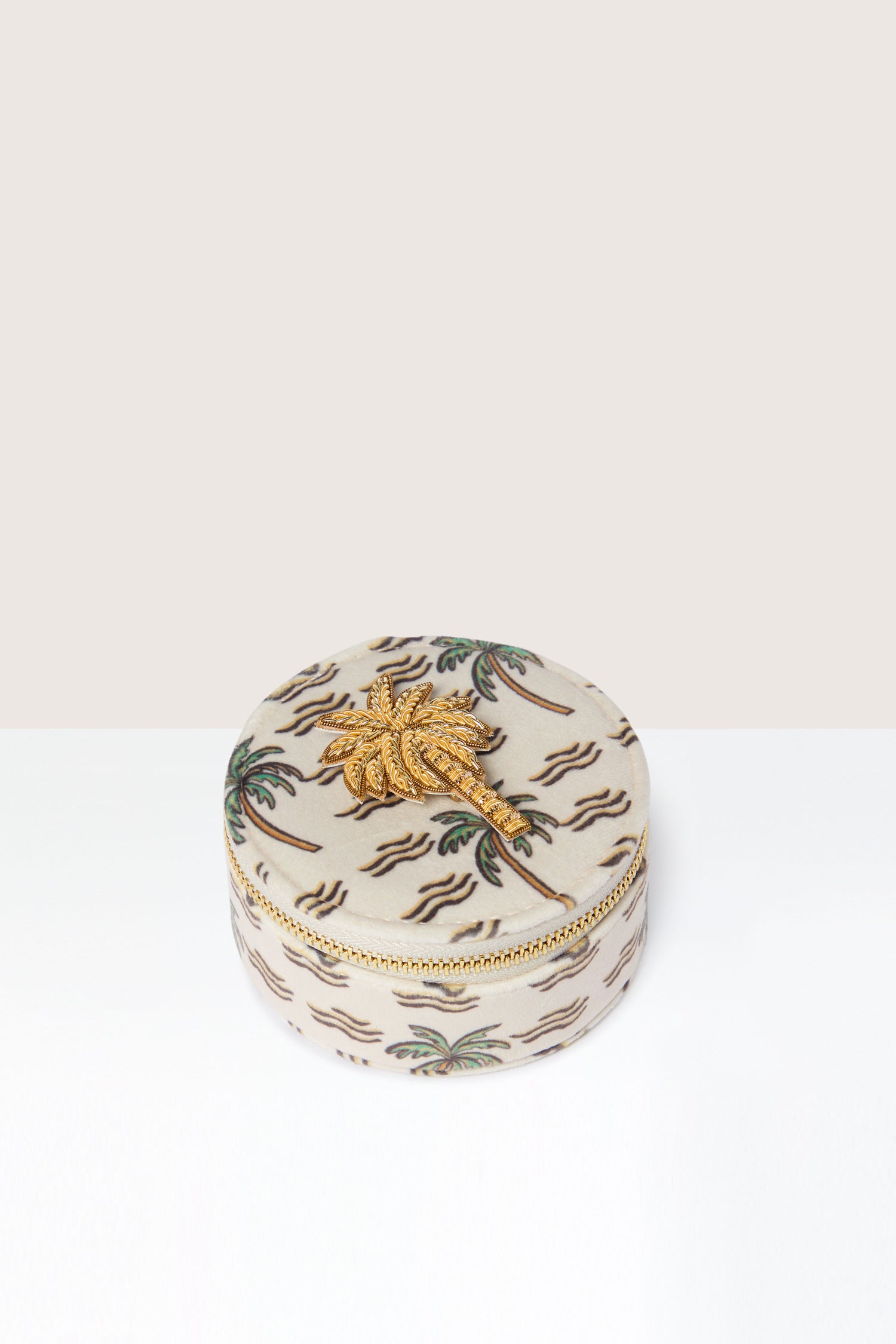 The Palm Jewellery Travel Pot is a round, fabric-covered accessory case with a zipper, adorned with a stunning gold palm tree design on the outside and complemented by a chic zebra print lining for added flair.