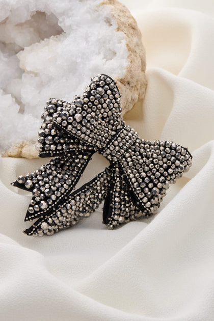 The Bow Brooch, adorned with studs, elegantly sits on a soft, cream-colored fabric backdrop with a mineral cluster in the background, making it a true statement piece.