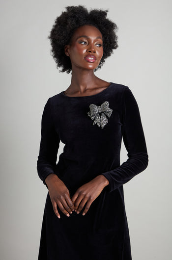 A person poses against a simple backdrop, radiating sophistication in a long-sleeved black dress accentuated by the Bow Brooch, a bold and eye-catching accessory on the chest. Their short curly hair complements their serene expression, enhancing the elegant ensemble.