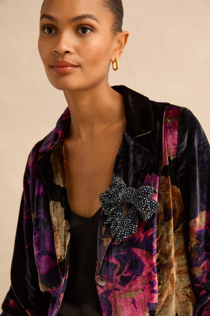 A large, dark Night Hand Beaded Bow Brooch pinned to the lapel of a floral velvet blazer.