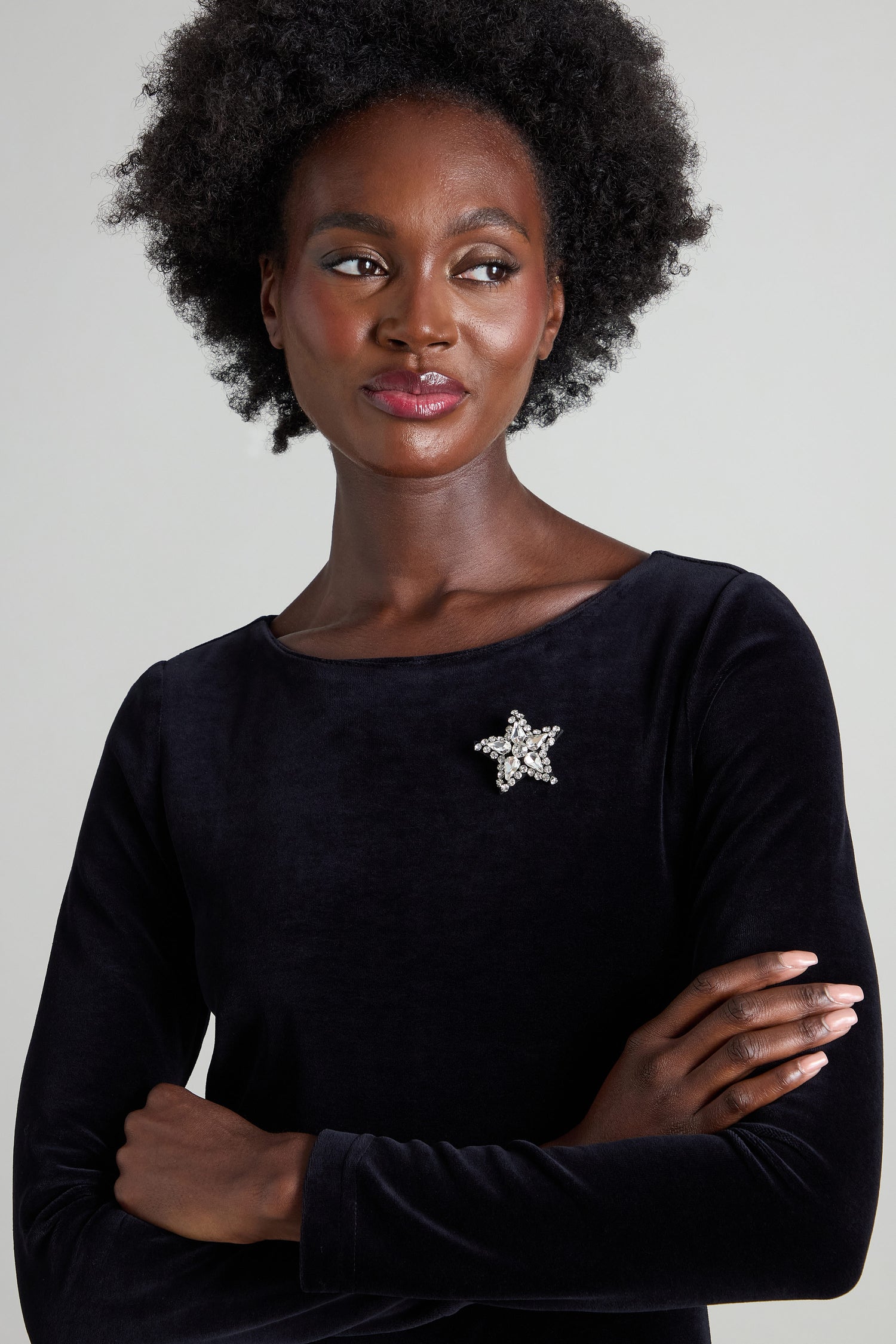 With curly hair, a person wears a dark velvet top featuring the Star Brooch, which is embellished with sparkling rhinestones. Their arms are crossed as they gaze off to the side.