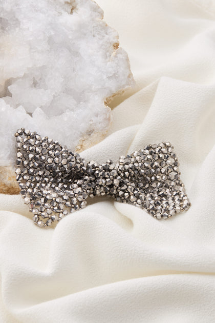 A Bow Tie Brooch with a metallic finish lies on a white fabric, providing an elegant shimmer next to a crystalline mineral rock.