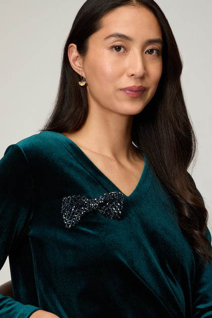 A woman with long dark hair wears a teal velvet top, gold earrings, and the Hand Beaded Bow Tie Brooch, looking at the camera against a neutral background.