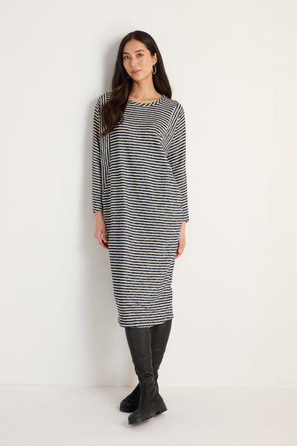 A woman stands against a white wall wearing the Textured Stripe Jersey Dress, which features black and white stripes, long sleeves, and is paired with black knee-high boots.