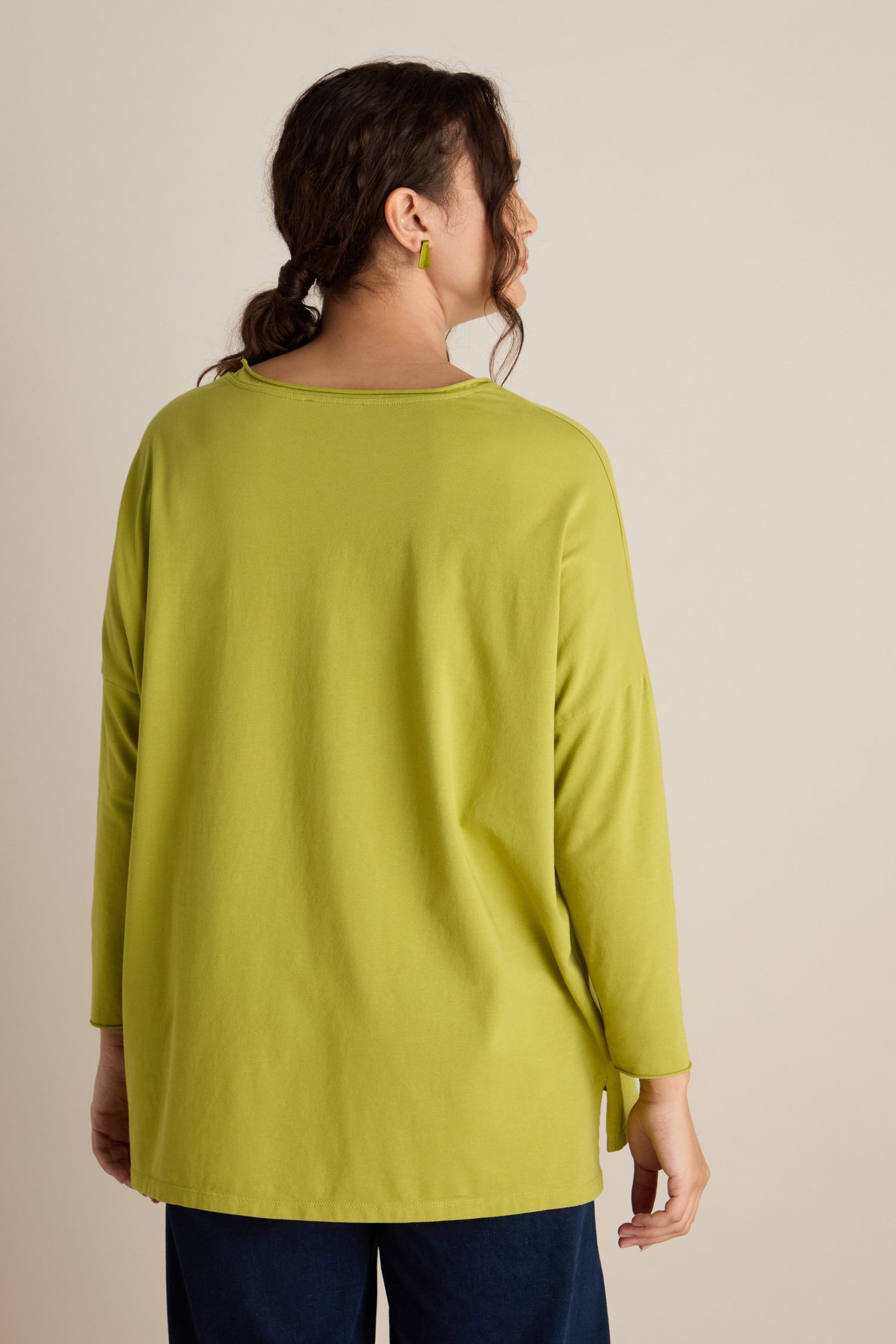 A person with dark hair tied back is seen from behind wearing the Cotton Jersey Relaxed Top in long-sleeve, lime-green and dark pants against a plain background, exuding a relaxed body posture that complements the subtle hues of the Autumn season.