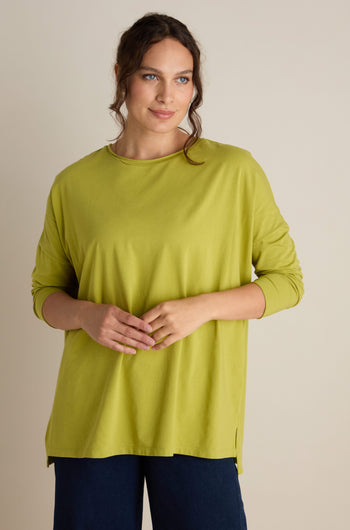 A person with long, wavy hair wears the loose, green Cotton Jersey Relaxed Top and dark pants, standing against a neutral background, perfectly embodying the relaxed vibes of the Autumn season.