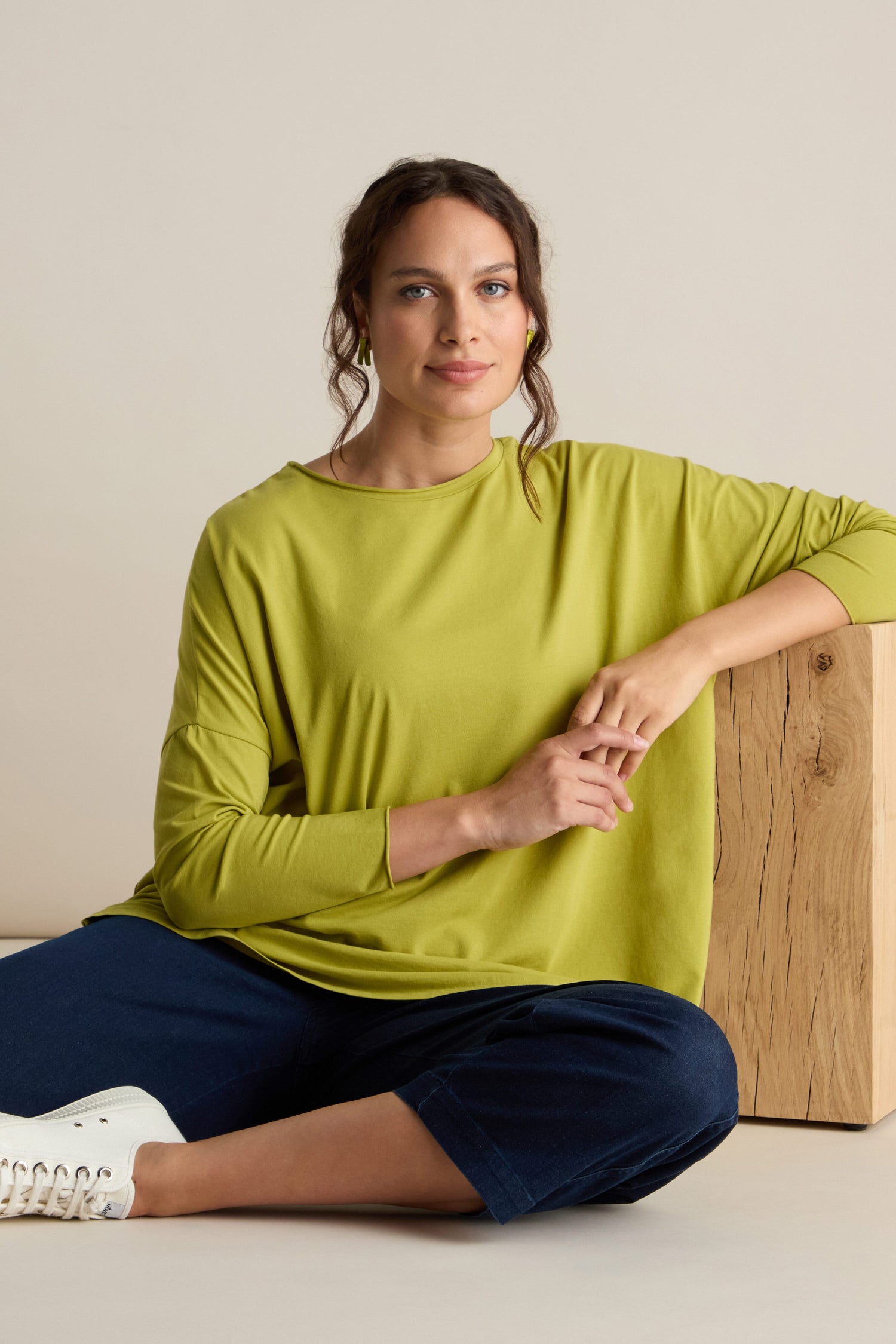 A person with a relaxed body sits on the floor with legs crossed, leaning against a wooden block. They are wearing a Cotton Jersey Relaxed Top in lime green, dark blue pants, and white sneakers, embodying the comfort of the Autumn season.