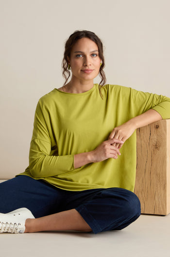 A person with a relaxed body sits on the floor with legs crossed, leaning against a wooden block. They are wearing a Cotton Jersey Relaxed Top in lime green, dark blue pants, and white sneakers, embodying the comfort of the Autumn season.