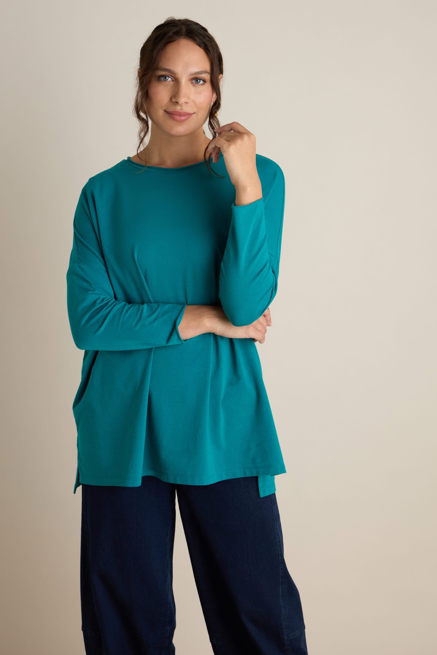 A person stands against a neutral background, wearing the Cotton Jersey Relaxed Top in teal and navy blue pants. They have long, wavy hair and a relaxed expression, with one hand touching their face. The relaxed body language perfectly captures the essence of the autumn season.