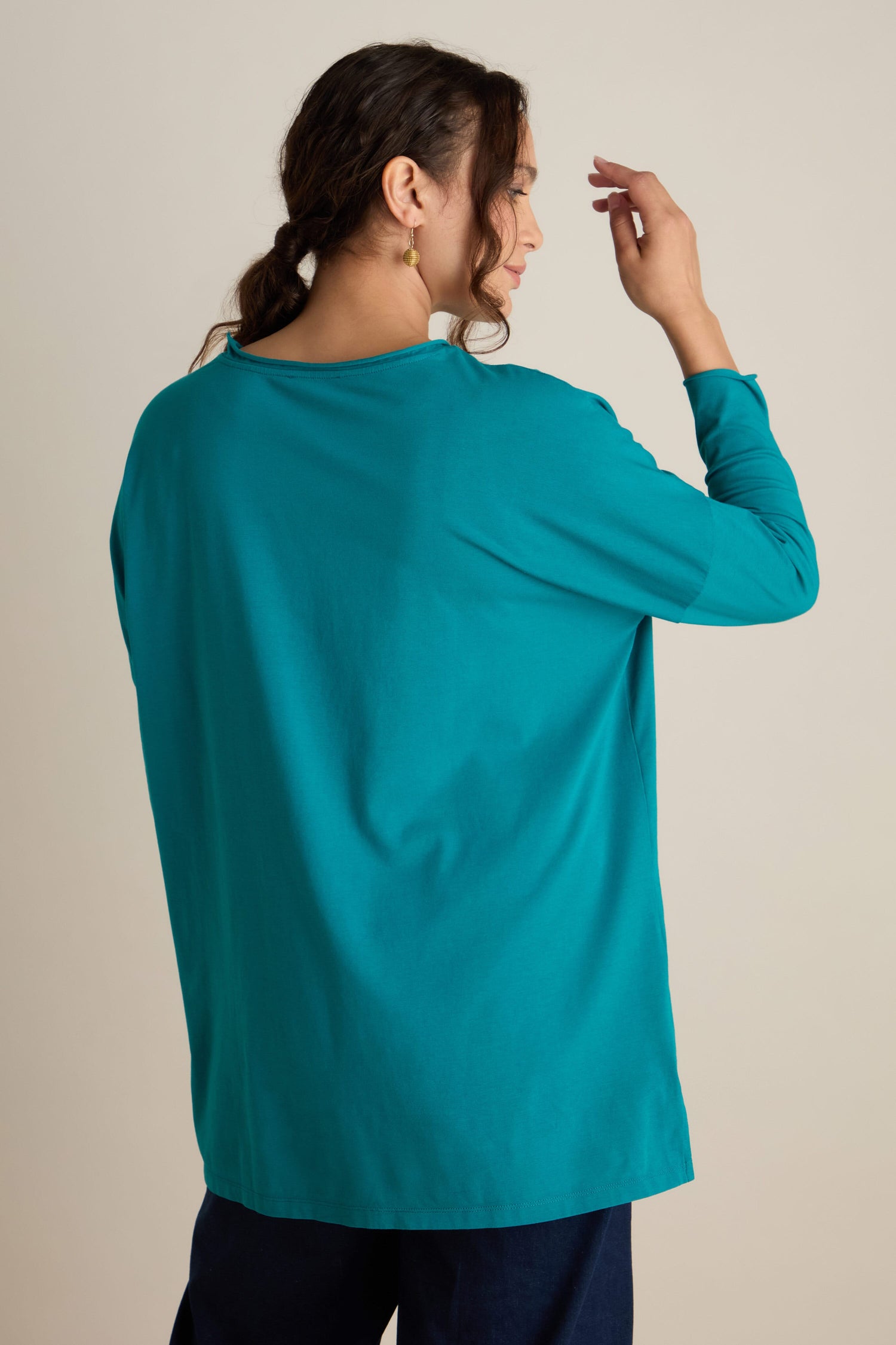 A person with long hair in a ponytail, dressed in a teal Cotton Jersey Relaxed Top, facing away, with a relaxed body and hand touching the back of the head, embodying the calm of the autumn season.