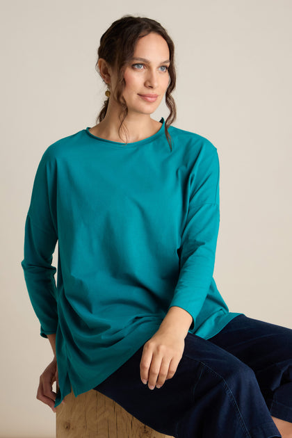 Person sitting on a wooden block wearing a Cotton Jersey Relaxed Top in teal and dark pants, with a relaxed body posture, looking slightly to the side with a neutral expression. The scene exudes the calm of the Autumn season.