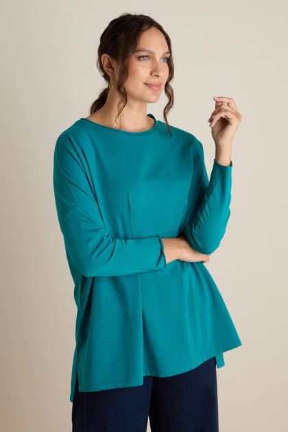 A woman wearing a Cotton Jersey Relaxed Top in teal stands against a plain background, looking off to the side while holding a finger near her face. With her long, curly hair tied back, she exudes a relaxed body posture that complements the serene feeling of the Autumn season.